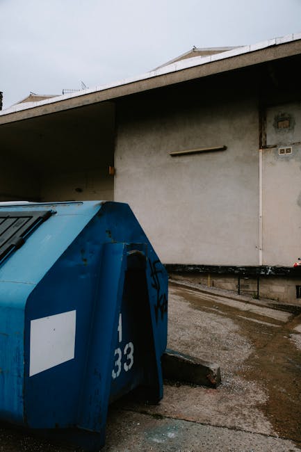 A large blue waste bin with a white identification label and partially visible graffiti, positioned on a concrete surface adjacent to a building with plain grayish exterior walls. The bin has a sloped